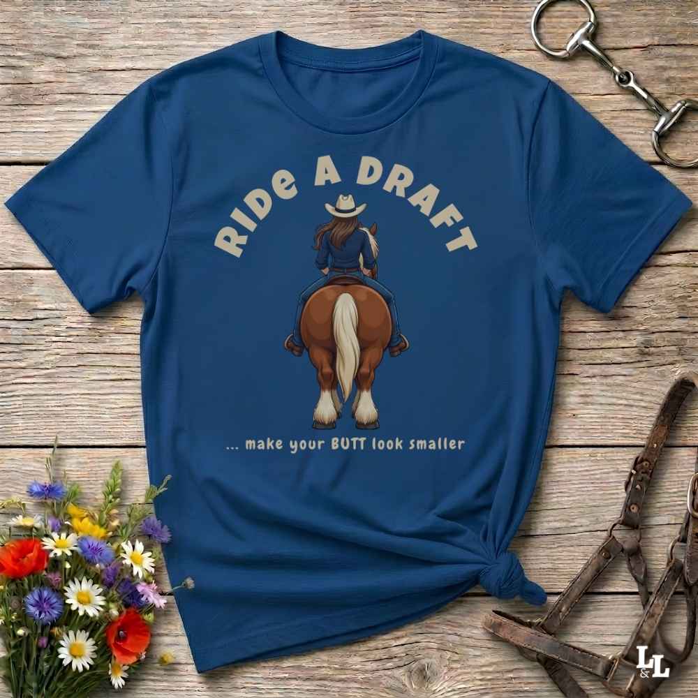A navy blue graphic t-shirt laid flat on a rustic wooden surface, styled with a small bouquet of wildflowers, a horse bit, and a leather halter. The shirt features a graphic of a cowboy riding a brown horse from behind, with the text 'RIDE A DRAFT .