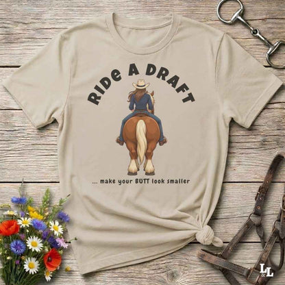A beige graphic t-shirt laid flat on a rustic wooden surface, styled with equestrian accessories and a small bouquet of wildflowers. The shirt features a graphic of a cowboy riding a horse with the text 'RIDE A DRAFT .