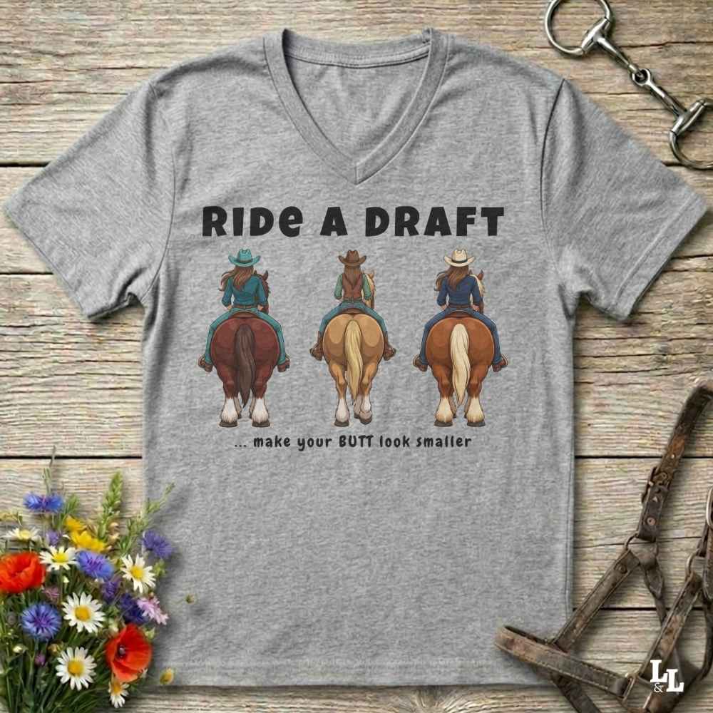 A heather gray V-neck t-shirt laid flat on a rustic wooden surface, featuring a graphic of three cowboys riding horses from behind with the text 'RIDE A DRAFT .