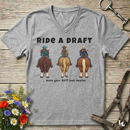 A heather gray V-neck t-shirt laid flat on a rustic wooden surface, featuring a graphic of three cowboys riding horses from behind with the text 'RIDE A DRAFT .