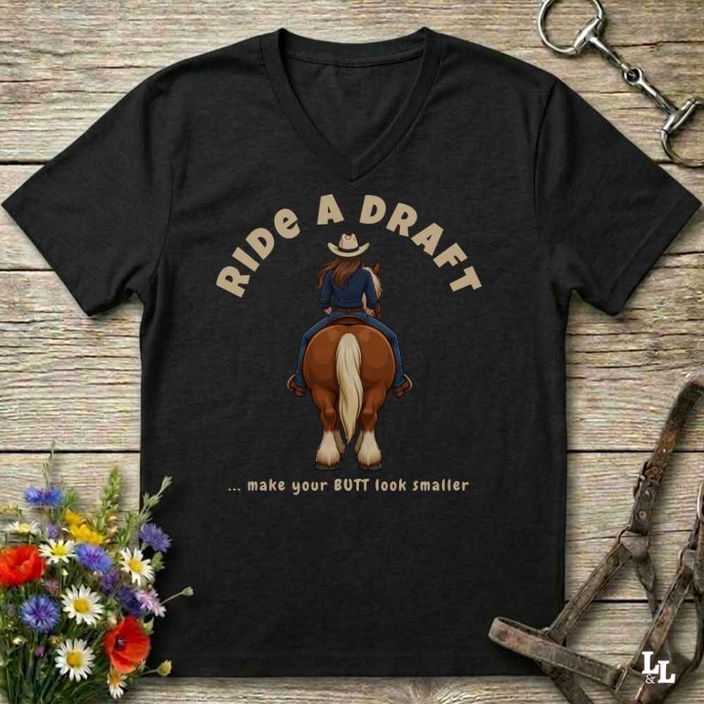 A black V-neck t-shirt laid flat on a rustic wooden surface, featuring a graphic of a cowboy riding a brown horse with the text 'RIDE A DRAFT .