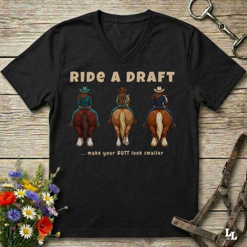 A black V-neck t-shirt laid flat on a rustic wooden surface, featuring a graphic of three cowboys riding horses with the text 'RIDE A DRAFT .