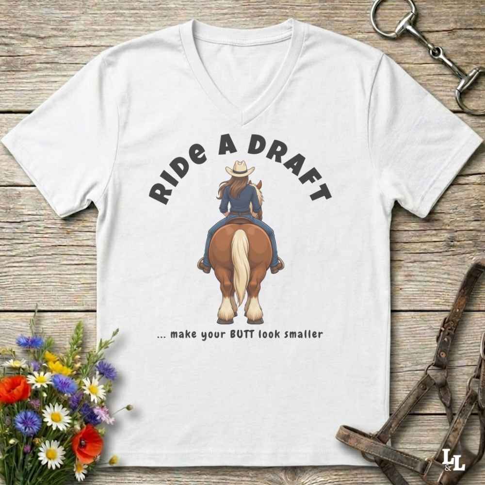 A white V-neck t-shirt laid flat on a rustic wooden surface, featuring a graphic of a woman riding a horse from behind with the text 'RIDE A DRAFT .