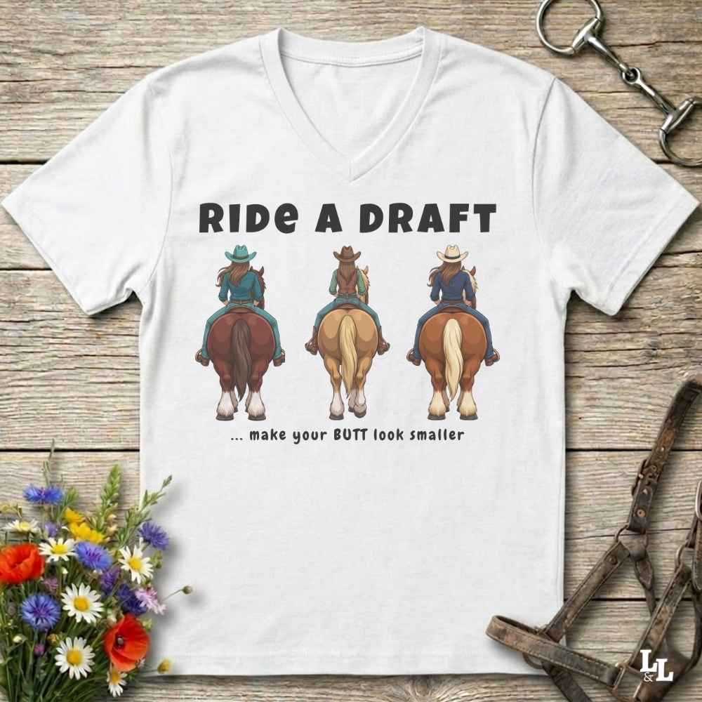 A white V-neck t-shirt laid flat on a rustic wooden surface, featuring a graphic of three cowboys riding horses from behind with the text 'RIDE A DRAFT .