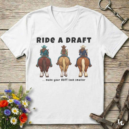 A white V-neck t-shirt laid flat on a rustic wooden surface, featuring a graphic of three cowboys riding horses from behind with the text 'RIDE A DRAFT .
