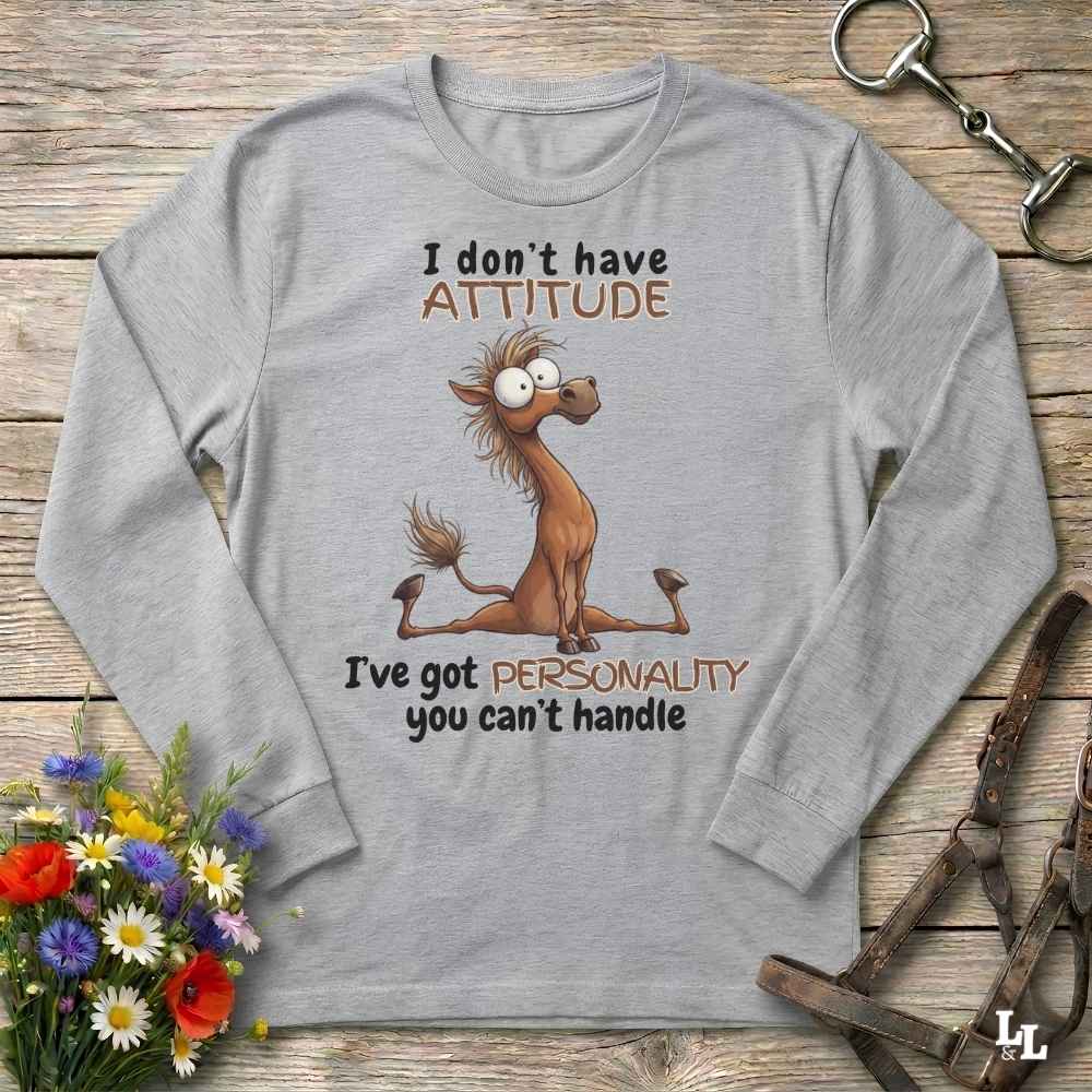 A light gray long-sleeve t-shirt with a cartoon horse graphic and the text 'I don't have ATTITUDE I've got PERSONALITY you can't handle', displayed on a rustic wooden background with flowers and leather straps.