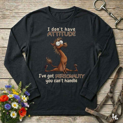 A black long-sleeve graphic t-shirt laid flat on a rustic wooden background, featuring a cartoon horse and the text 'I don't have ATTITUDE I've got PERSONALITY you can't handle'. The shirt is styled with horse tack and a small flower bouquet.