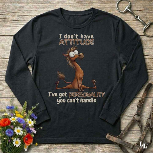 A black long-sleeve graphic t-shirt laid flat on a rustic wooden background, featuring a cartoon horse and the text 'I don't have ATTITUDE I've got PERSONALITY you can't handle'. The shirt is styled with horse tack and a small flower bouquet.