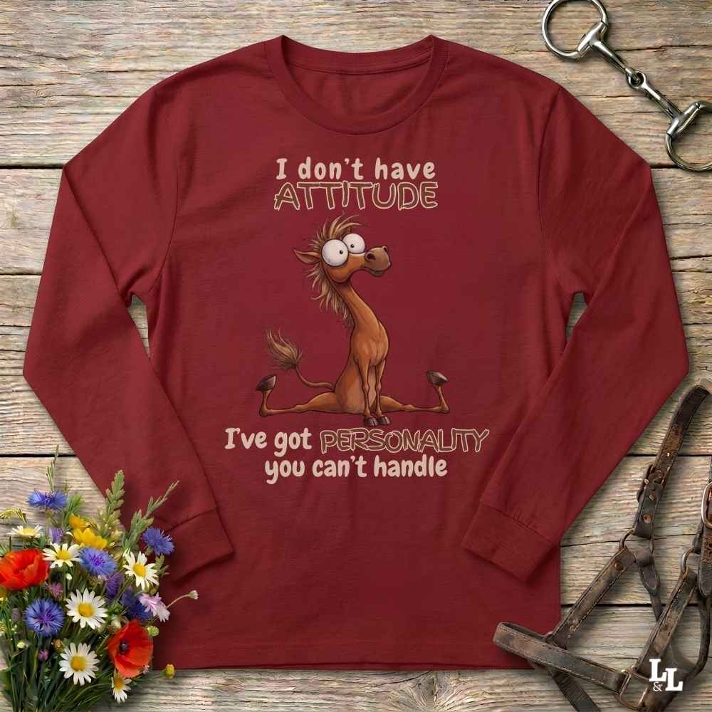 A long-sleeve crewneck t-shirt in maroon red, featuring a cartoon illustration of a nervous donkey with the text 'I don't have ATTITUDE I've got PERSONALITY you can't handle'. The shirt is displayed flat on a rustic wooden background with decorative props including a flower bouquet and leather tack.