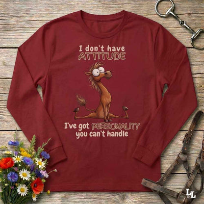 A long-sleeve crewneck t-shirt in maroon red, featuring a cartoon illustration of a nervous donkey with the text 'I don't have ATTITUDE I've got PERSONALITY you can't handle'. The shirt is displayed flat on a rustic wooden background with decorative props including a flower bouquet and leather tack.