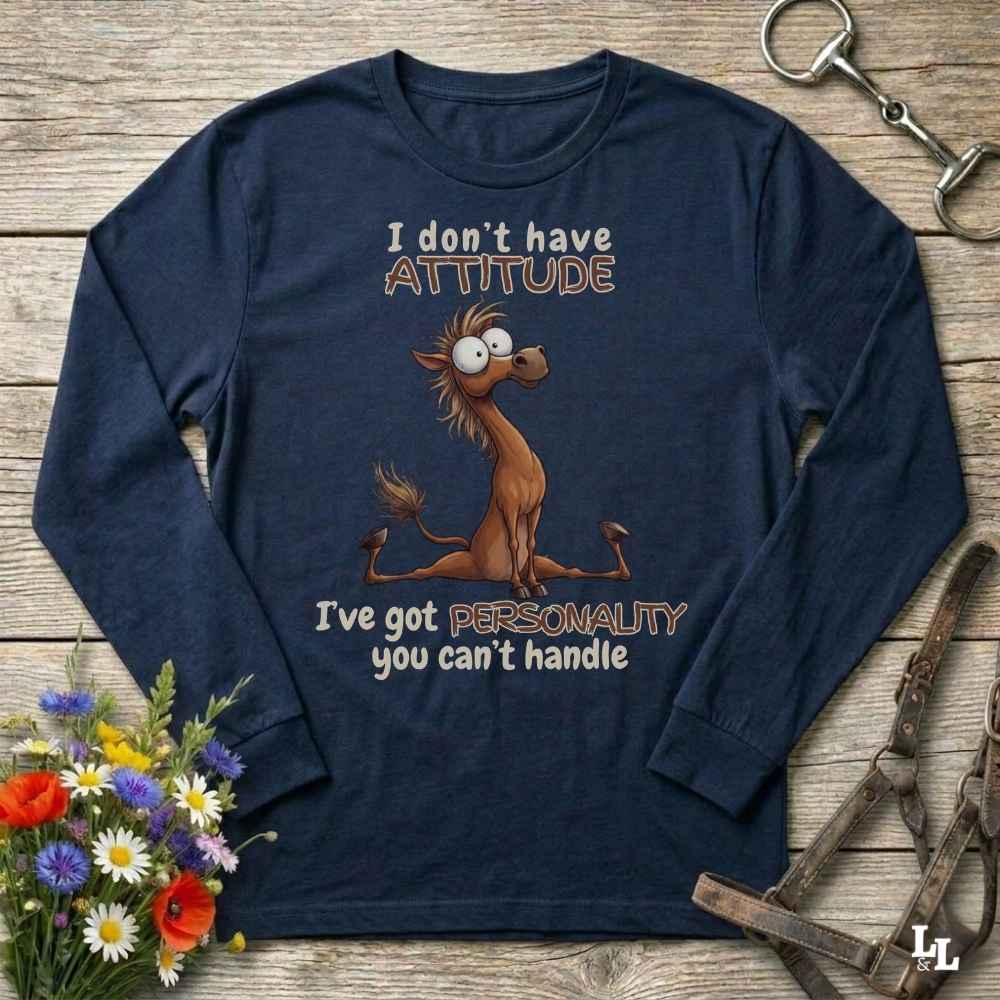 A navy blue long-sleeve t-shirt with a cartoon horse graphic and the text 'I don't have ATTITUDE I've got PERSONALITY you can't handle', laid flat on a rustic wooden background with decorative flowers and horse tack.