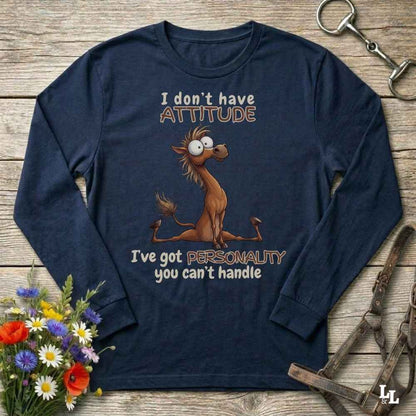 A navy blue long-sleeve t-shirt with a cartoon horse graphic and the text 'I don't have ATTITUDE I've got PERSONALITY you can't handle', laid flat on a rustic wooden background with decorative flowers and horse tack.