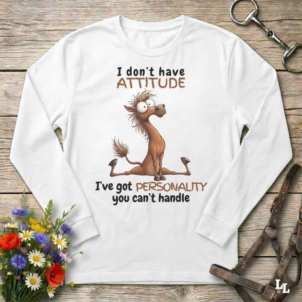 A white long-sleeve t-shirt with a humorous graphic of a cartoon horse and the text 'I don't have ATTITUDE I've got PERSONALITY you can't handle', displayed on a rustic wooden background with equestrian accessories and flowers.
