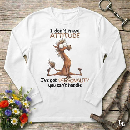 A white long-sleeve t-shirt with a humorous graphic of a cartoon horse and the text 'I don't have ATTITUDE I've got PERSONALITY you can't handle', displayed on a rustic wooden background with equestrian accessories and flowers.