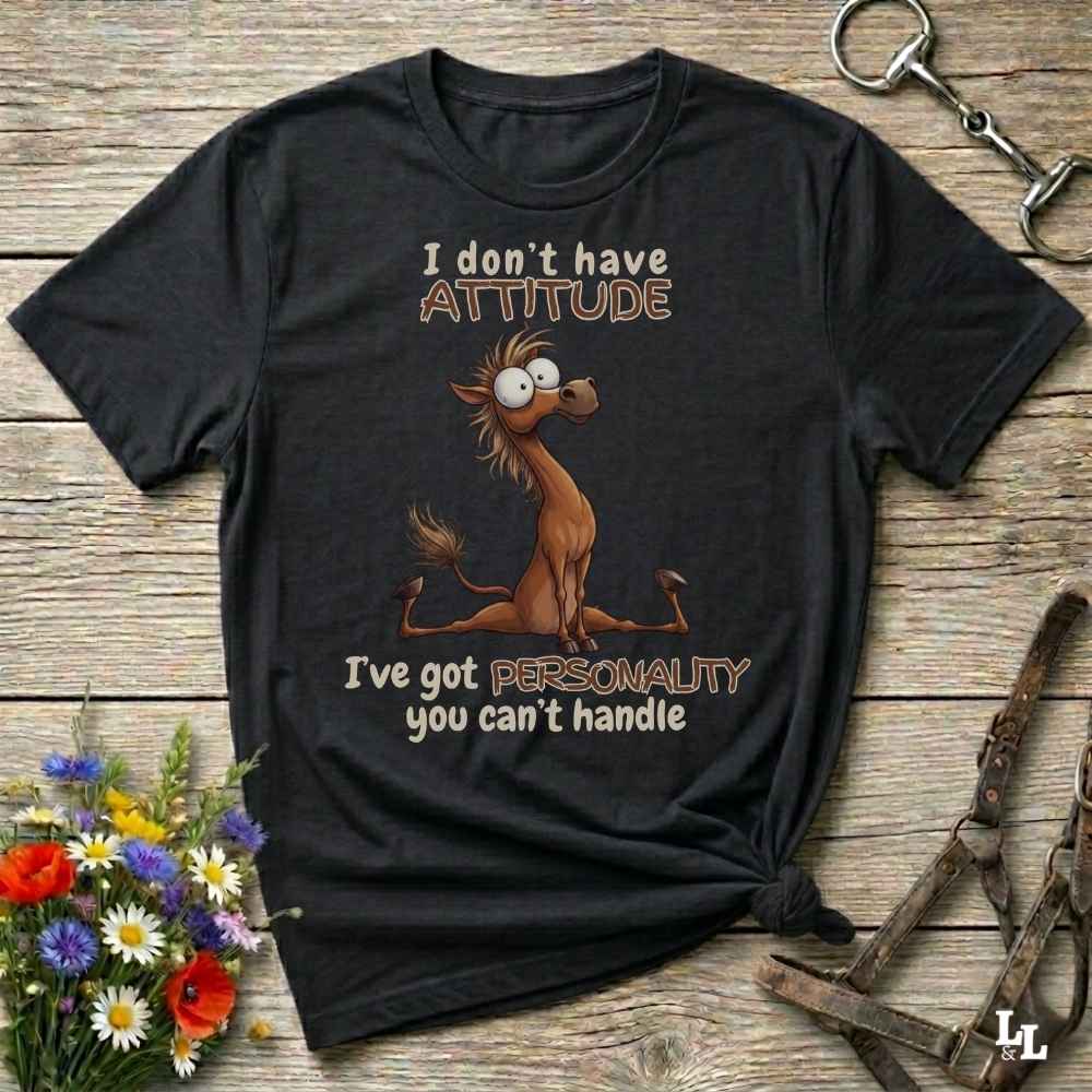 A black graphic t-shirt featuring a cartoon horse and the text 'I don't have ATTITUDE I've got PERSONALITY you can't handle', displayed on a rustic wooden background with decorative flowers and horse tack.
