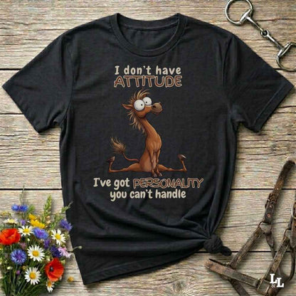 A black graphic t-shirt featuring a cartoon horse and the text 'I don't have ATTITUDE I've got PERSONALITY you can't handle', displayed on a rustic wooden background with decorative flowers and horse tack.