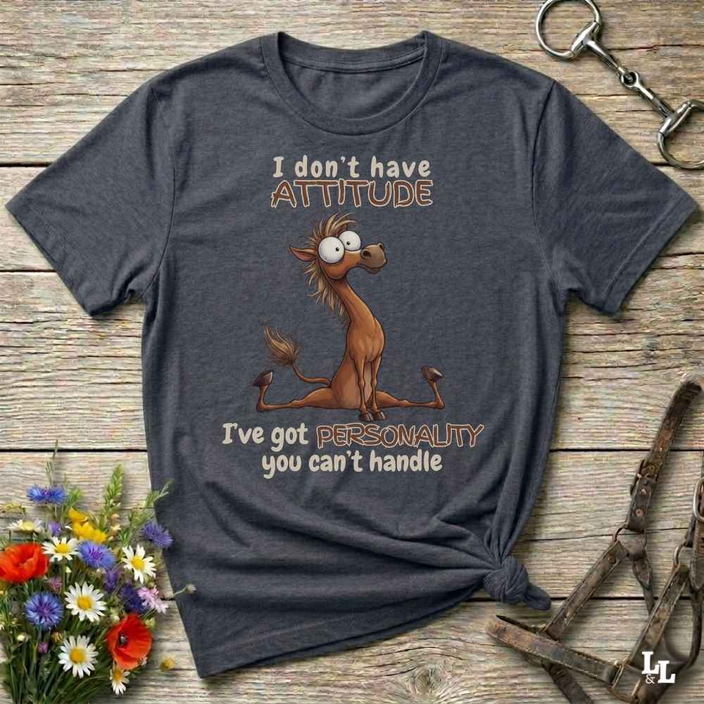 A dark gray graphic t-shirt laid flat on a rustic wooden surface, styled with a small bouquet of flowers, a horse bit, and a leather bridle. The t-shirt features a cartoon donkey graphic with the text 'I don't have ATTITUDE I've got PERSONALITY you can't handle'.