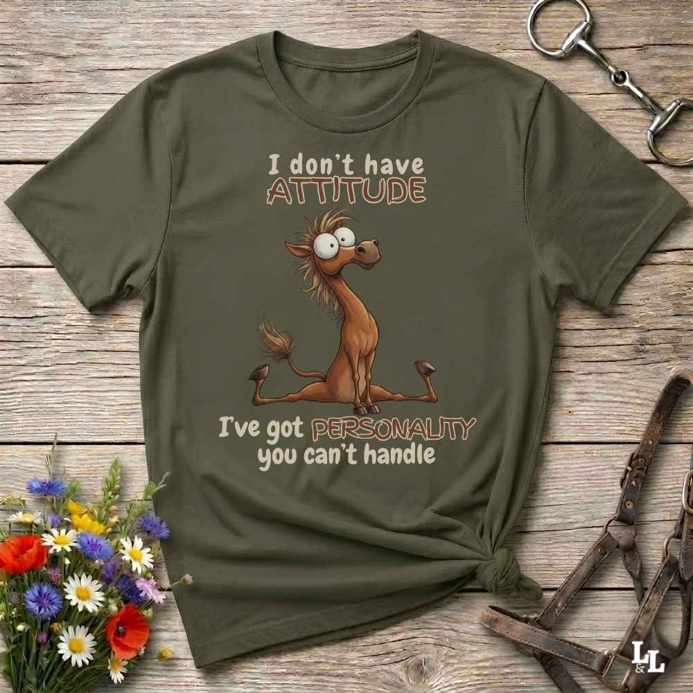 An olive green graphic t-shirt laid flat on a rustic wooden background, styled with a horse bit in the top right and a small bouquet of colorful flowers in the bottom left. The t-shirt features a cartoon donkey graphic with the text 'I don't have ATTITUDE I've got PERSONALITY you can't handle'.