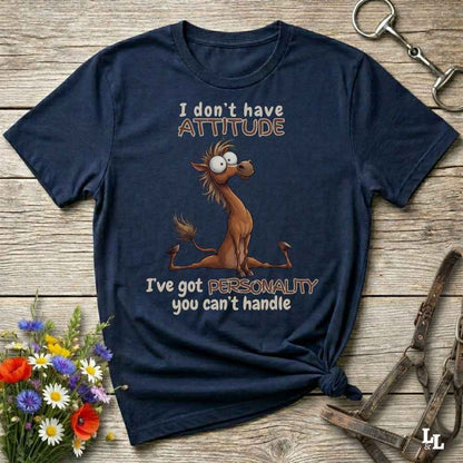 A navy blue graphic t-shirt featuring a cartoon camel and the text 'I don't have ATTITUDE I've got PERSONALITY you can't handle', laid flat on a rustic wooden background with decorative flowers and horse tack.
