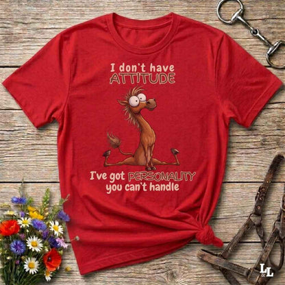 A red graphic t-shirt featuring a cartoon donkey and the text 'I don't have ATTITUDE I've got PERSONALITY you can't handle', displayed on a rustic wooden background with decorative flowers and leather tack.