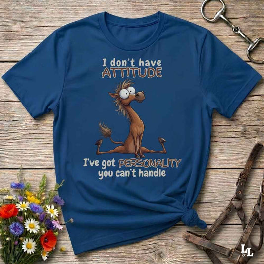 A navy blue graphic t-shirt laid flat on a rustic wooden surface, styled with horse tack and a small bouquet of flowers. The shirt features a cartoon donkey and the text 'I don't have ATTITUDE I've got PERSONALITY you can't handle'.
