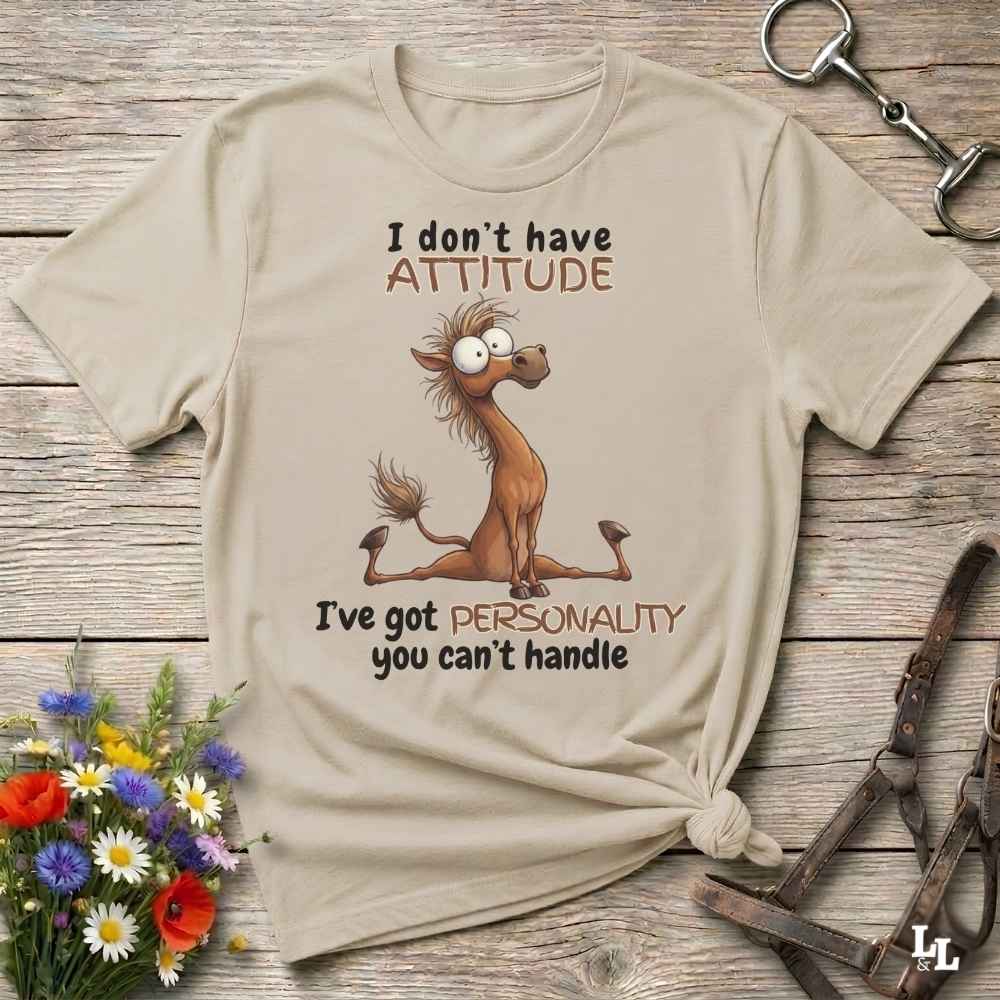 A beige graphic t-shirt laid flat on a rustic wooden surface, featuring a cartoon brown horse and the text 'I don't have ATTITUDE I've got PERSONALITY you can't handle'. The shirt is styled with equestrian accessories and small flowers.