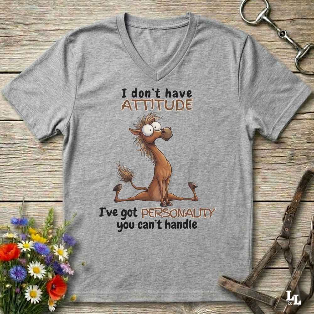 A heather gray V-neck t-shirt with a cartoon horse graphic and the text 'I don't have ATTITUDE I've got PERSONALITY you can't handle', laid flat on a rustic wooden surface with flowers and horse tack props.