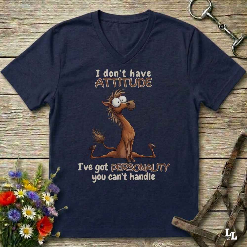 A navy blue V-neck t-shirt with a cartoon horse graphic and the text 'I don't have ATTITUDE I've got PERSONALITY you can't handle', displayed on a rustic wooden background with equestrian accessories and flowers.