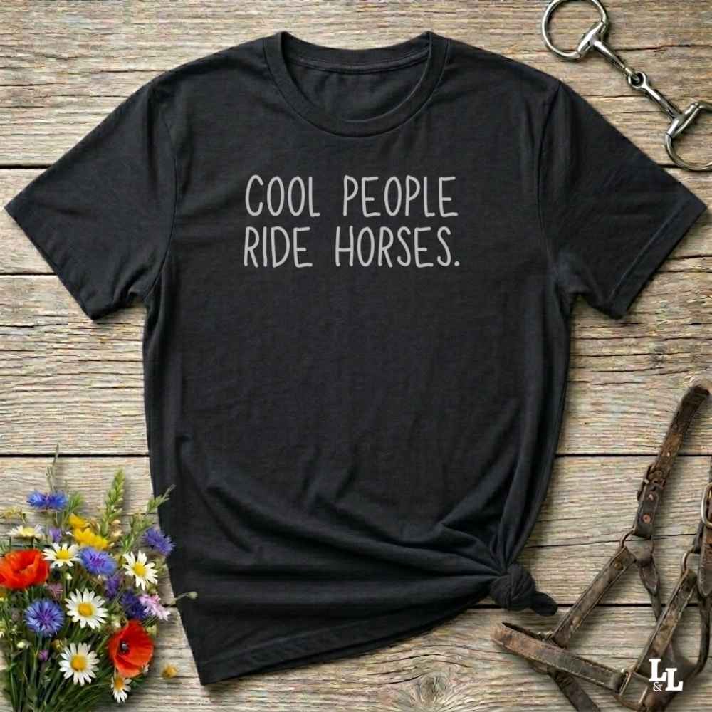A black short-sleeve t-shirt with the phrase 'COOL PEOPLE RIDE HORSES.' printed in white across the chest, styled flat on a rustic wooden surface with equestrian accessories and flowers.