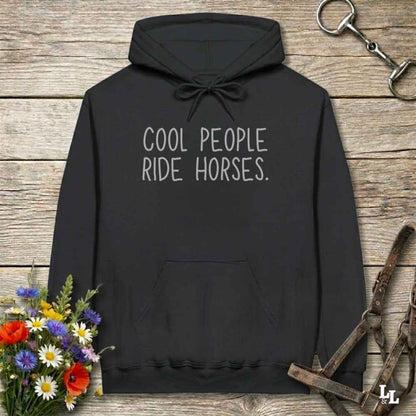 A black pullover hoodie laid flat on a rustic wood surface, with white text reading “COOL PEOPLE RIDE HORSES.” on the chest.