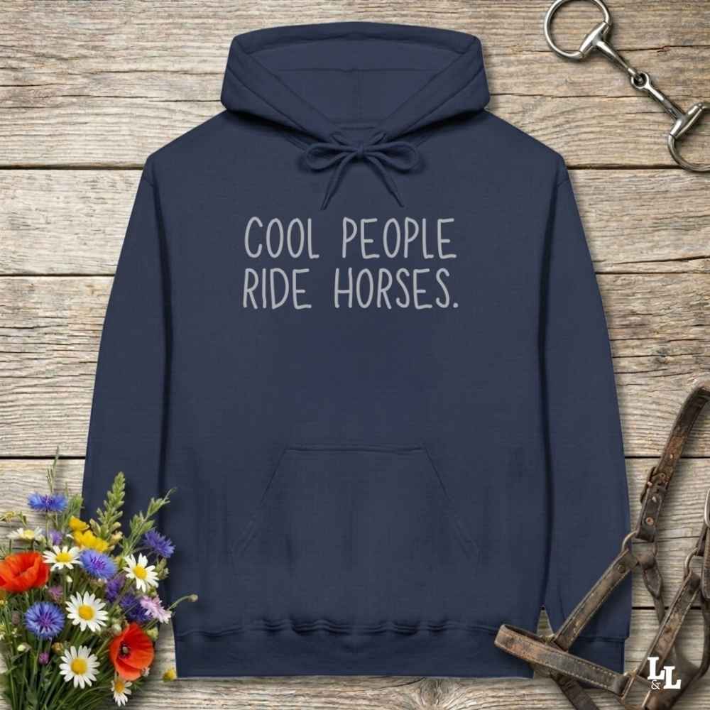 A navy blue pullover hoodie with the phrase 'COOL PEOPLE RIDE HORSES.' printed in white across the chest, displayed on a rustic wooden background with equestrian-themed props including a horse bit, bridle strap, and a small bouquet of flowers.