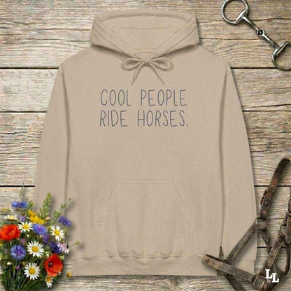 A beige pullover hoodie with the text 'COOL PEOPLE RIDE HORSES.' printed in dark gray on the chest, displayed on a rustic wooden background with equestrian-themed props including a halter, a horse bit, and a small bouquet of wildflowers.