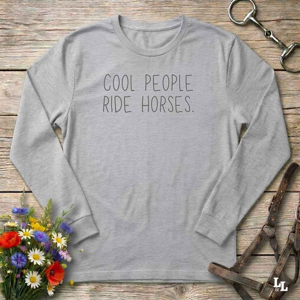 A light gray long-sleeve t-shirt with the text 'COOL PEOPLE RIDE HORSES.' printed in dark gray on the chest, styled in a flat-lay on a rustic wooden background with equestrian accessories and flowers.