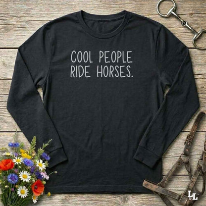 A black long-sleeve t-shirt with the text 'COOL PEOPLE RIDE HORSES.' printed in white on the chest, laid flat on a rustic wooden background with equestrian accessories and flowers.