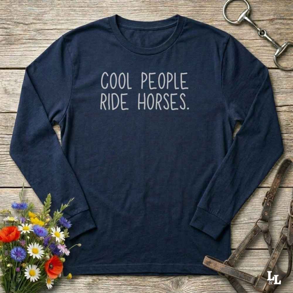 A navy blue long-sleeve t-shirt with the phrase 'COOL PEOPLE RIDE HORSES.' printed in white across the chest, styled in a flat-lay on a rustic wooden background with equestrian and floral props.