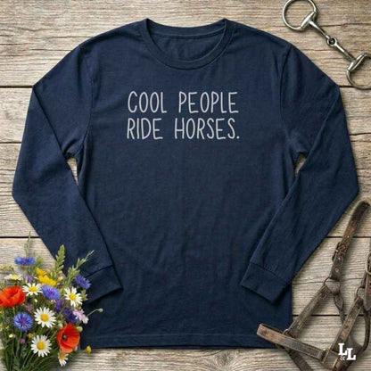 A navy blue long-sleeve t-shirt with the phrase 'COOL PEOPLE RIDE HORSES.' printed in white across the chest, styled in a flat-lay on a rustic wooden background with equestrian and floral props.