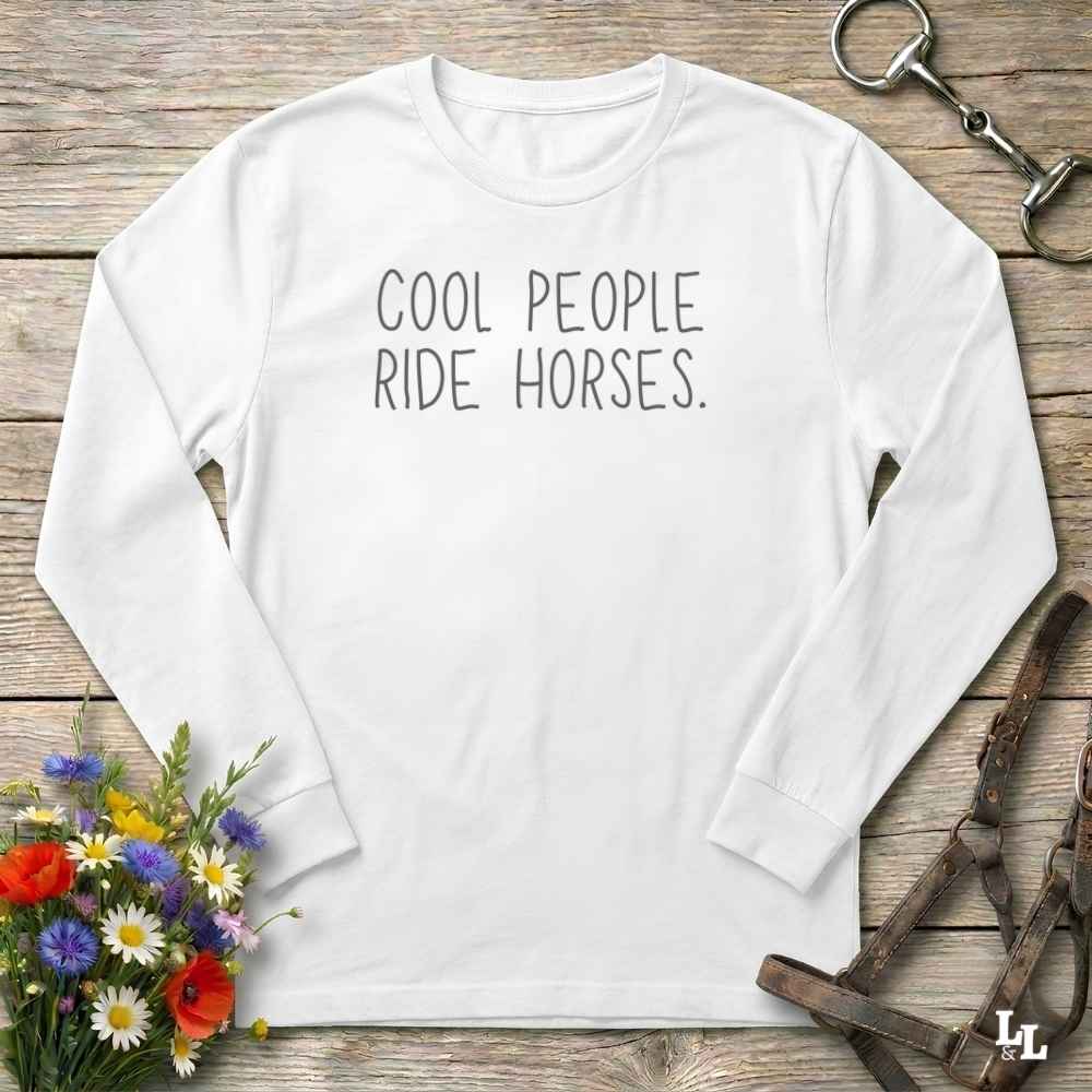 A white long-sleeve t-shirt with the text 'COOL PEOPLE RIDE HORSES.' printed in black across the chest, displayed in a flat-lay arrangement on a rustic wooden background with equestrian and floral props.