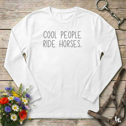 A white long-sleeve t-shirt with the text 'COOL PEOPLE RIDE HORSES.' printed in black across the chest, displayed in a flat-lay arrangement on a rustic wooden background with equestrian and floral props.
