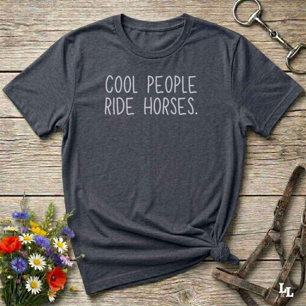 A dark charcoal gray t-shirt with the text 'COOL PEOPLE RIDE HORSES.' printed in white on the front, styled in a flat-lay on a rustic wooden background with equestrian accessories and flowers.