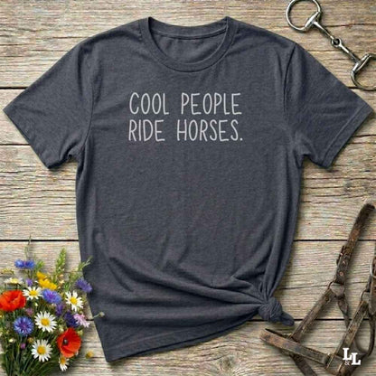 A dark charcoal gray t-shirt with the text 'COOL PEOPLE RIDE HORSES.' printed in white on the front, styled in a flat-lay on a rustic wooden background with equestrian accessories and flowers.