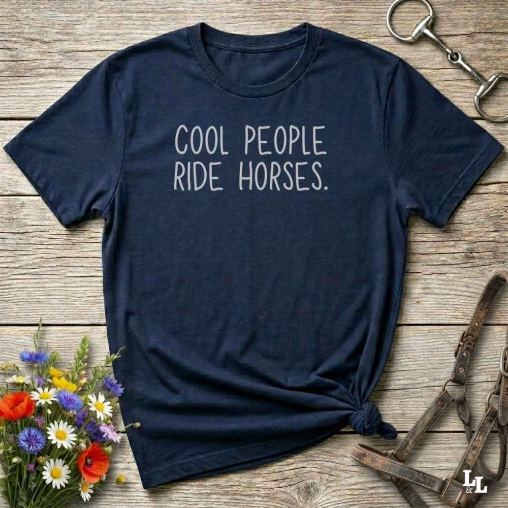 A navy blue graphic t-shirt with the text 'COOL PEOPLE RIDE HORSES.' printed in white on the chest, styled in a flat-lay on a rustic wooden background with equestrian and floral props.