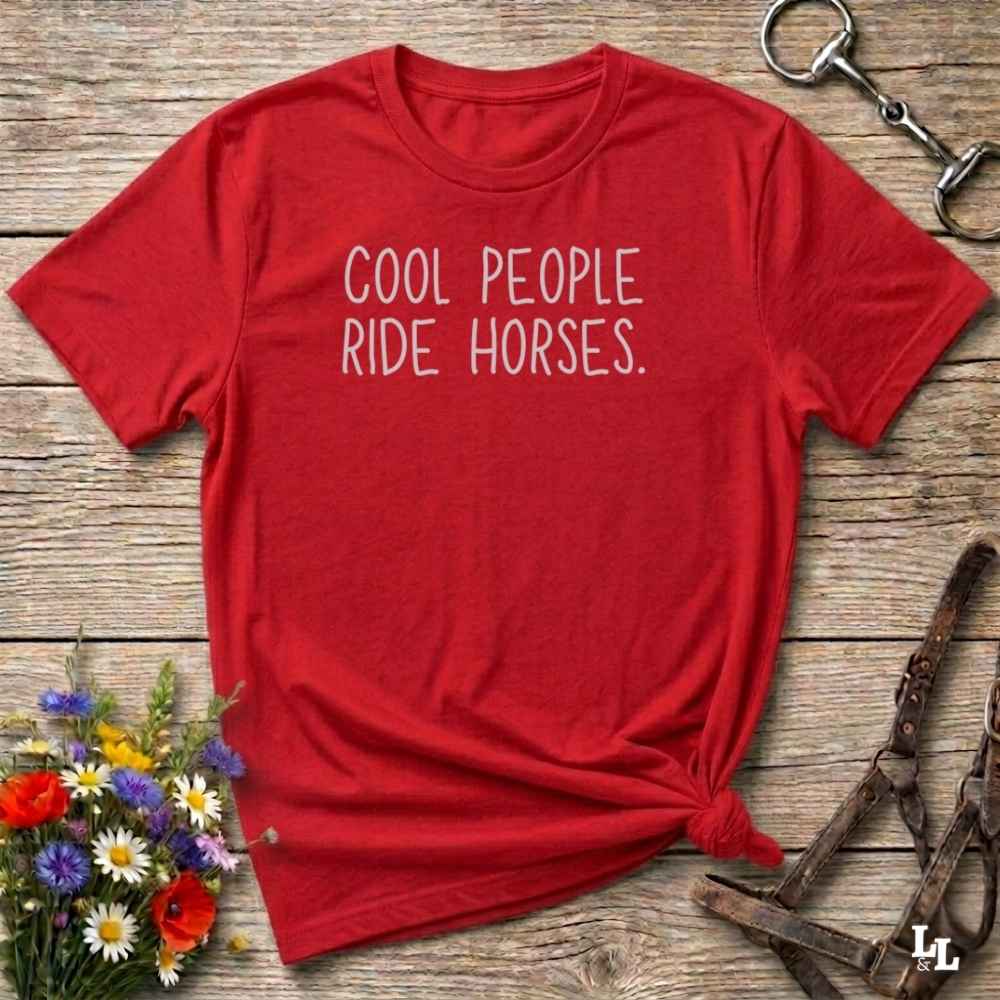 A red short-sleeve t-shirt with the phrase 'COOL PEOPLE RIDE HORSES.' printed in white across the chest, styled flat on a rustic wooden surface with flowers, a horse bit, and a leather bridle nearby.