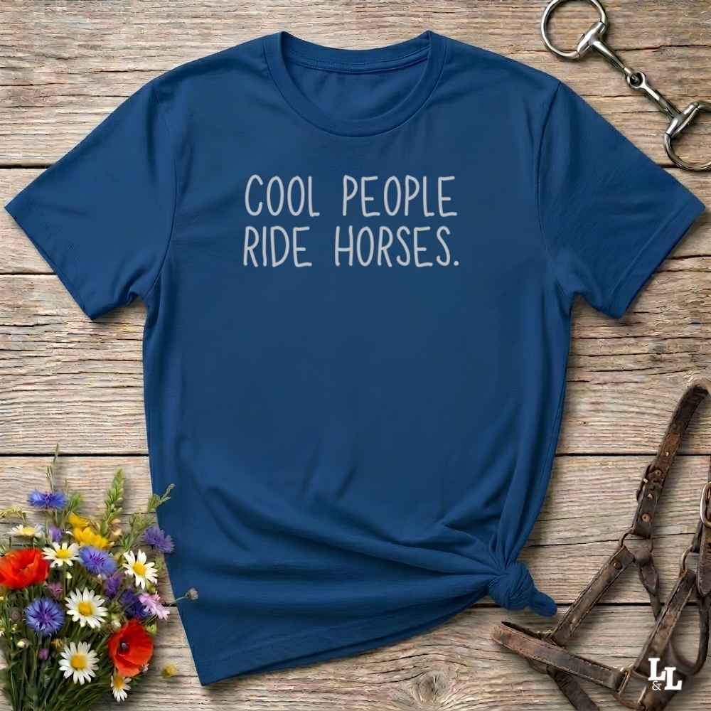 A navy blue short-sleeve t-shirt with the phrase 'COOL PEOPLE RIDE HORSES.' printed in white across the chest, styled in a flat-lay on a rustic wooden surface with equestrian accessories and flowers.
