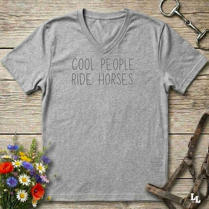 A heather gray V-neck t-shirt with the text 'COOL PEOPLE RIDE HORSES.' printed in black across the chest, displayed in a flat-lay arrangement on a rustic wooden background with equestrian accessories and flowers.