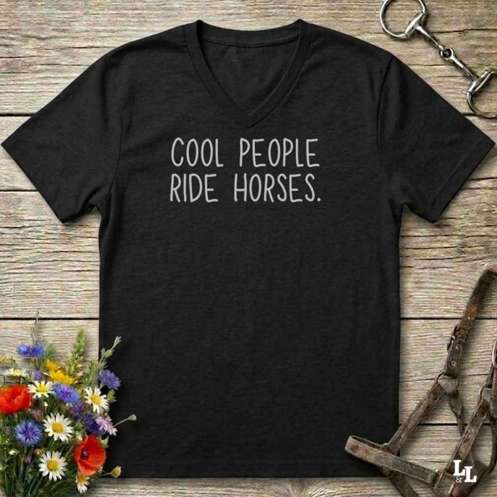 A black V-neck t-shirt with the text 'COOL PEOPLE RIDE HORSES.' printed in white on the chest, displayed on a rustic wooden background with equestrian and floral props.