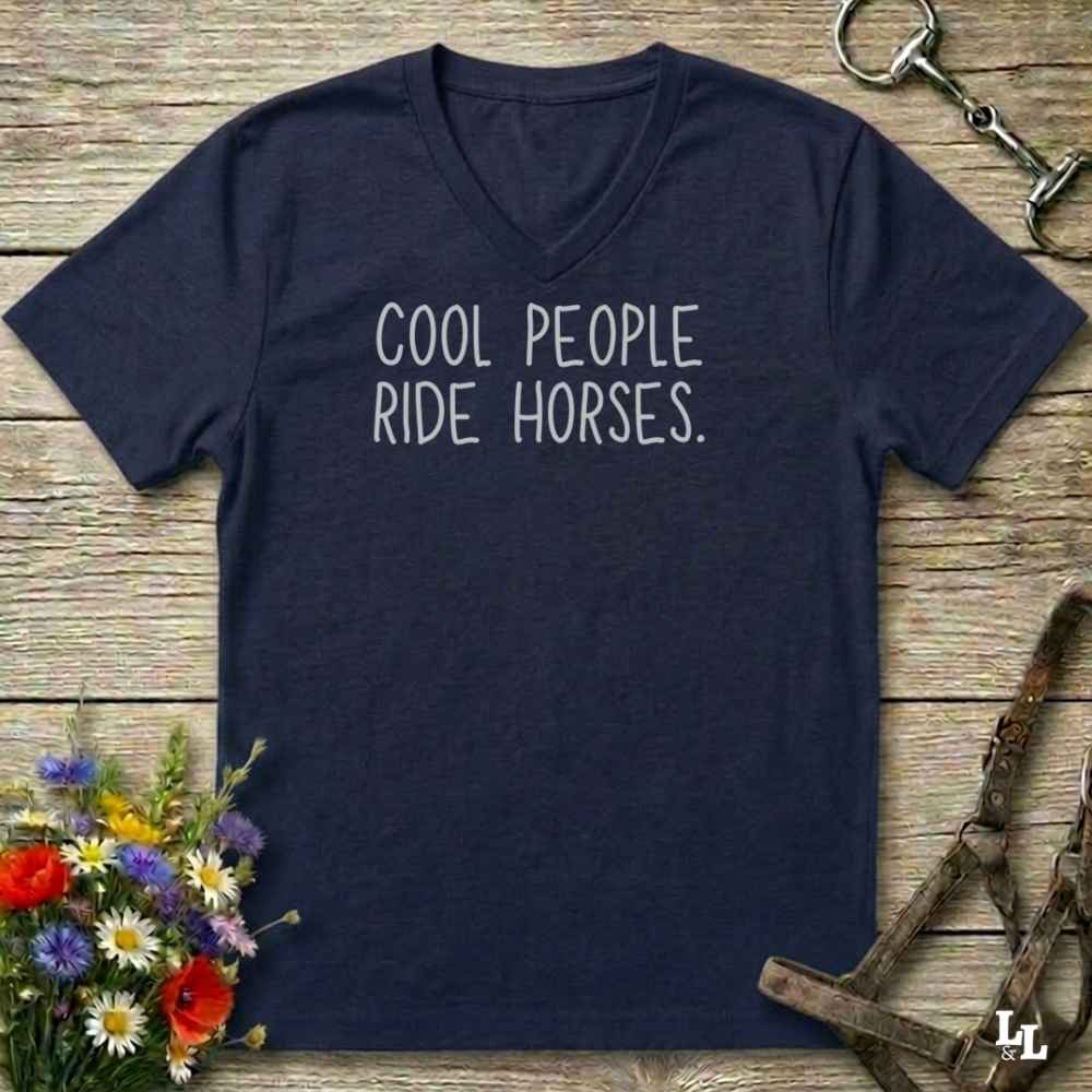 A navy blue V-neck t-shirt with the phrase 'COOL PEOPLE RIDE HORSES.' printed in white across the chest, laid flat on a rustic wooden surface with equestrian-themed props including a halter, horse bit, and wildflowers.