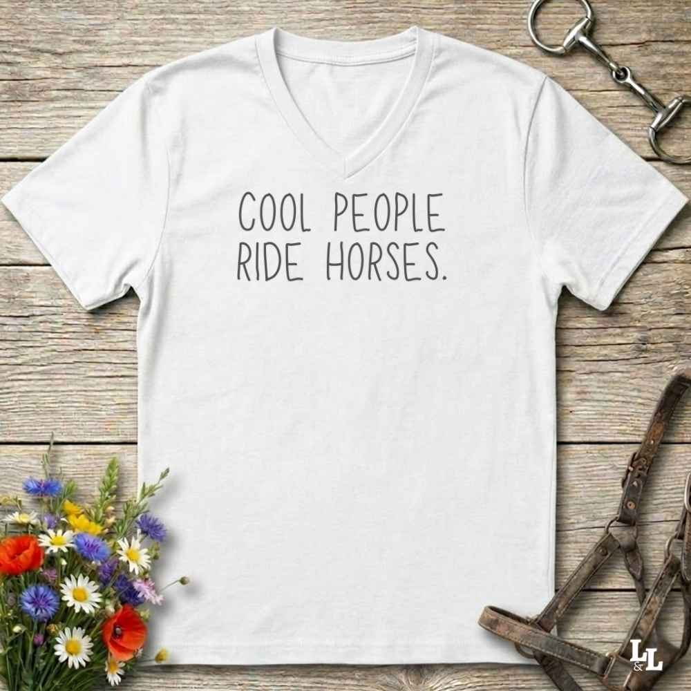 A white v-neck t-shirt with the text 'COOL PEOPLE RIDE HORSES.' printed in black across the chest, laid flat on a rustic wooden background with equestrian-themed props.