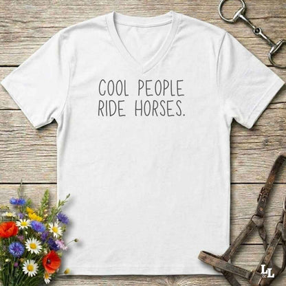 A white v-neck t-shirt with the text 'COOL PEOPLE RIDE HORSES.' printed in black across the chest, laid flat on a rustic wooden background with equestrian-themed props.