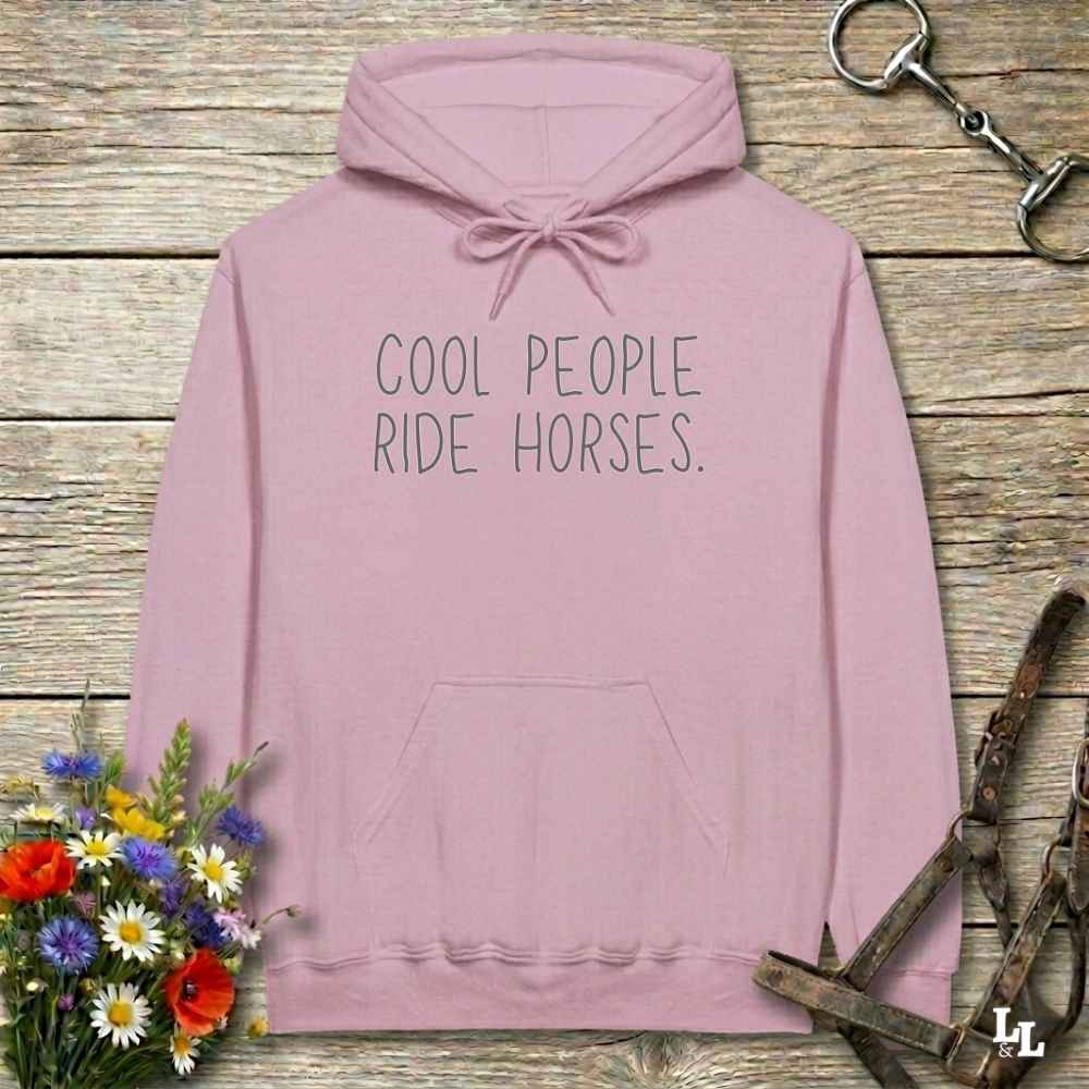 A dusty pink pullover hoodie with the text 'COOL PEOPLE RIDE HORSES.' printed in black on the chest, displayed on a rustic wooden background with equestrian-themed props including a horse bit, leather bridle, and a bouquet of wildflowers.