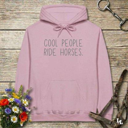 A dusty pink pullover hoodie with the text 'COOL PEOPLE RIDE HORSES.' printed in black on the chest, displayed on a rustic wooden background with equestrian-themed props including a horse bit, leather bridle, and a bouquet of wildflowers.